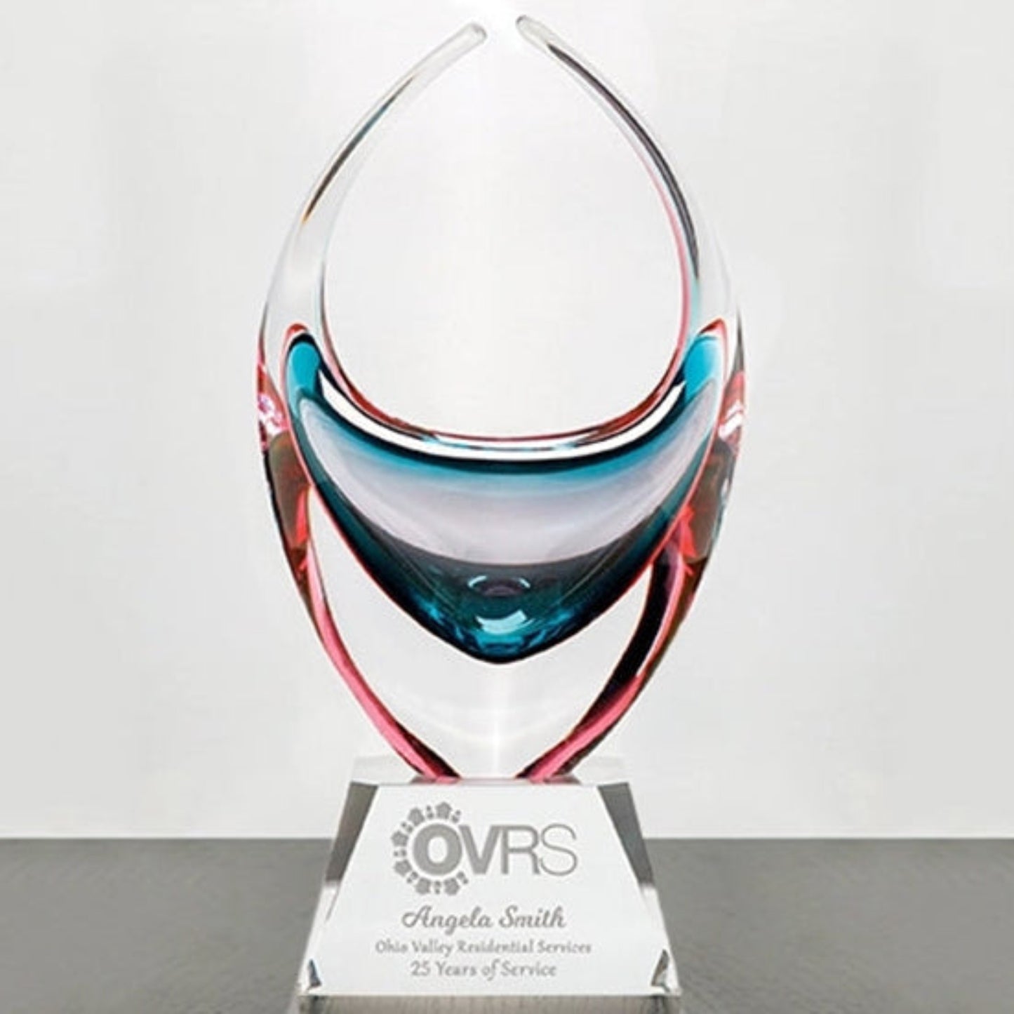 A clear glass award trophy with sculpted curved shapes in blue, pink, and red tones, mounted on a clear acrylic base with a printed plaque reading OVRS and Angela Smith.