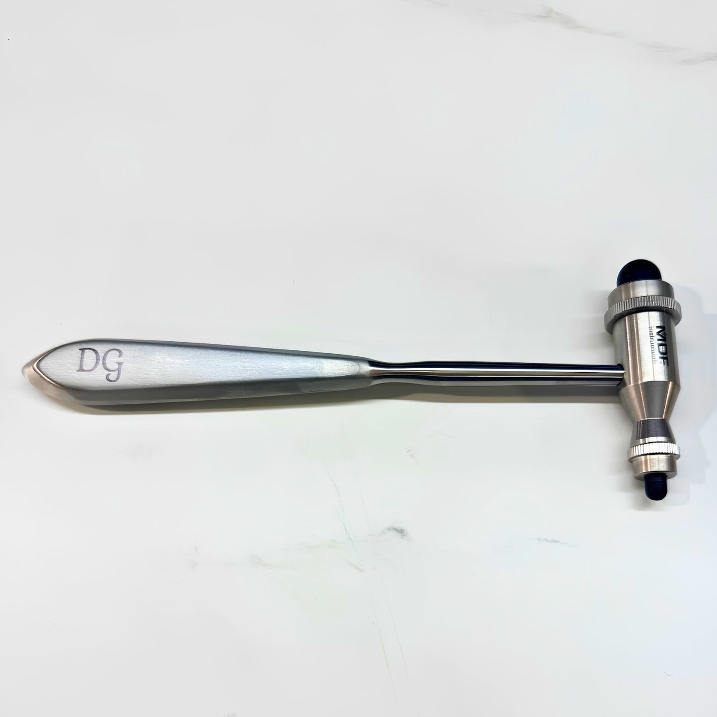 A metal reflex hammer with a polished silver handle engraved with 'DG' and a matching metal head with a black rubber tip, placed on a light gray marble surface.