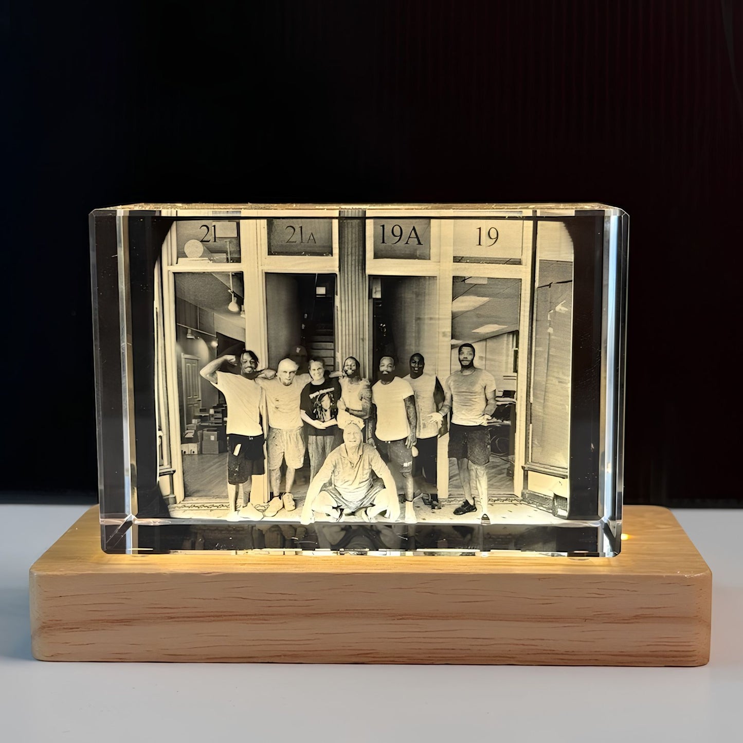 A custom rectangular clear crystal photo block displaying a black-and-white group photograph, mounted on a light-colored wooden base against a dark background.
