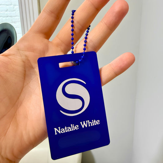 A hand holding a royal blue plastic key tag with a matching blue beaded chain. The tag features a white stylized 'S' logo and the text 'Natalie White'.