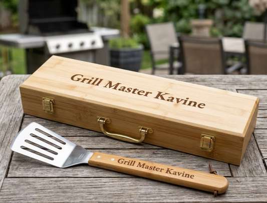 A personalized wooden gift box and matching spatula set engraved with 'Grill Master Kayine', displayed on a rustic wooden table with an outdoor patio grill in the background.