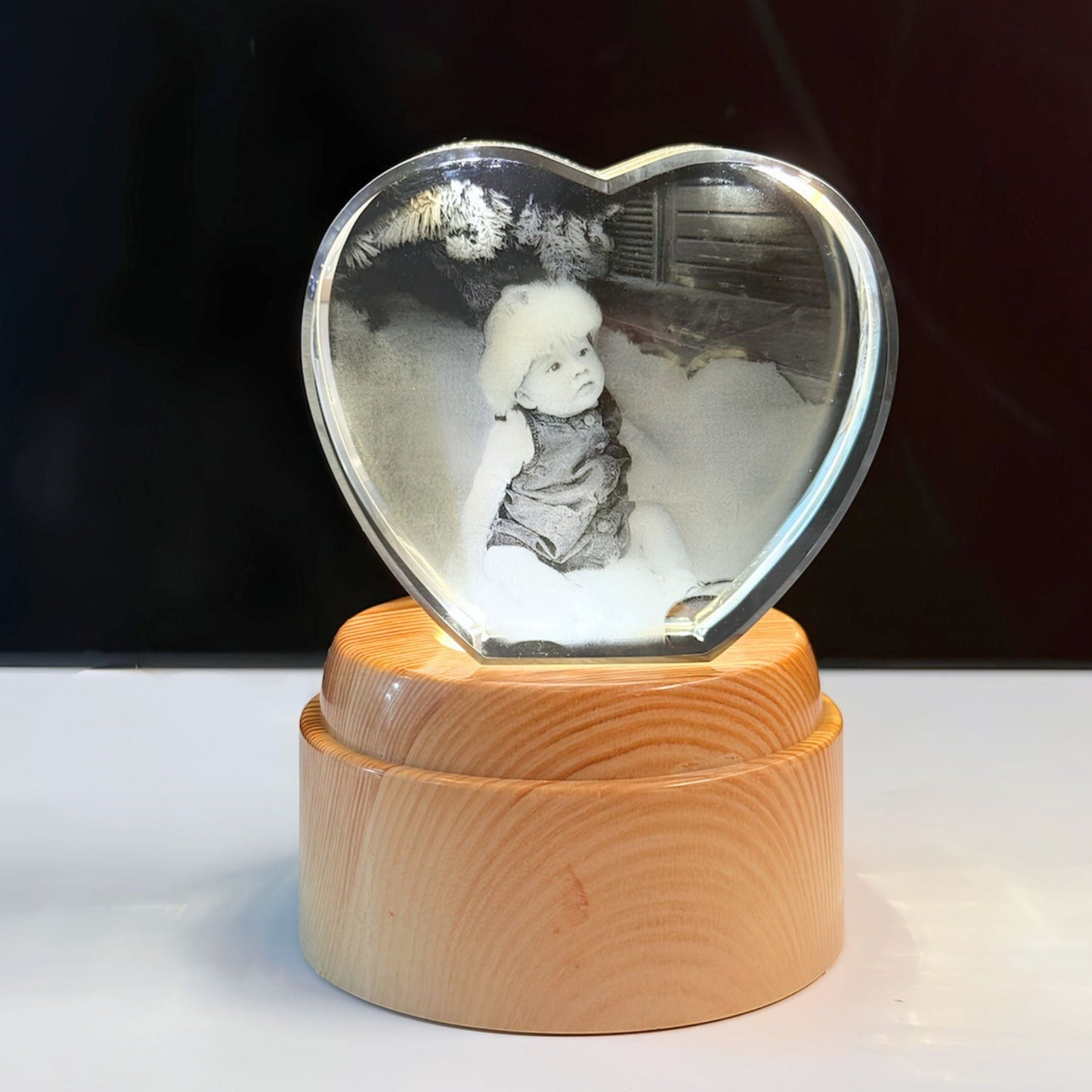 A heart-shaped clear crystal keepsake photo display resting on a round light wood base, showing a black-and-white photo of a child inside.