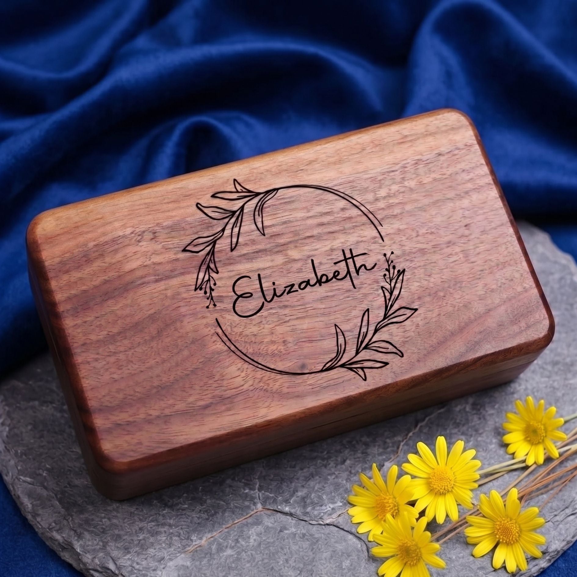 A rectangular wooden keepsake box with rounded corners, featuring an engraved floral wreath and the name 'Elizabeth' on the lid. The box is placed on a gray slate board with yellow daisies and draped dark blue fabric in the background.