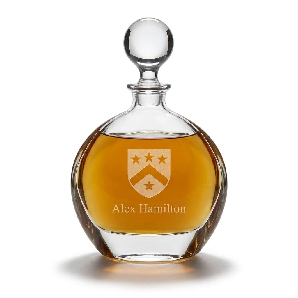 A personalized clear glass decanter with a round stopper, filled with amber liquor, featuring an engraved crest and the name 'Alex Hamilton' on the front, set against a plain white background.