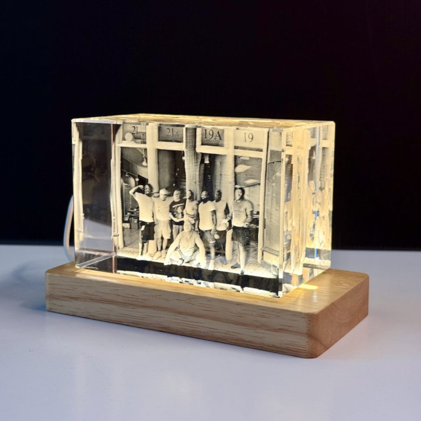 A custom acrylic photo box lamp with a warm white LED light glowing from within, showcasing an engraved black and white photograph of a group of people. The clear acrylic box sits on a rectangular light brown wooden base against a dark background.