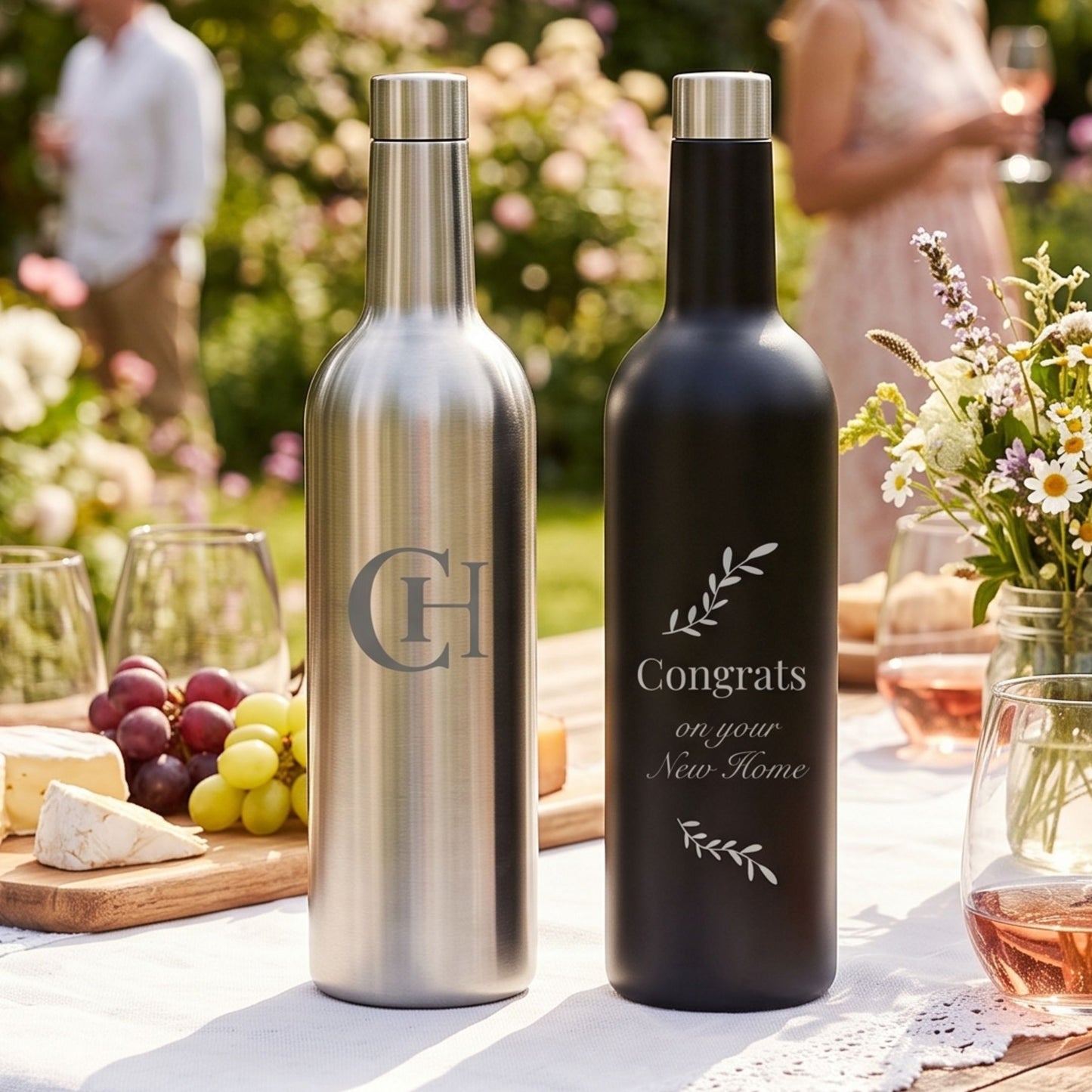 Two personalized wine bottle flasks on a white tablecloth at an outdoor garden party. One bottle is brushed silver with a gray monogram 'CH', and the other is matte black with a silver 'Congrats on your New Home' print.