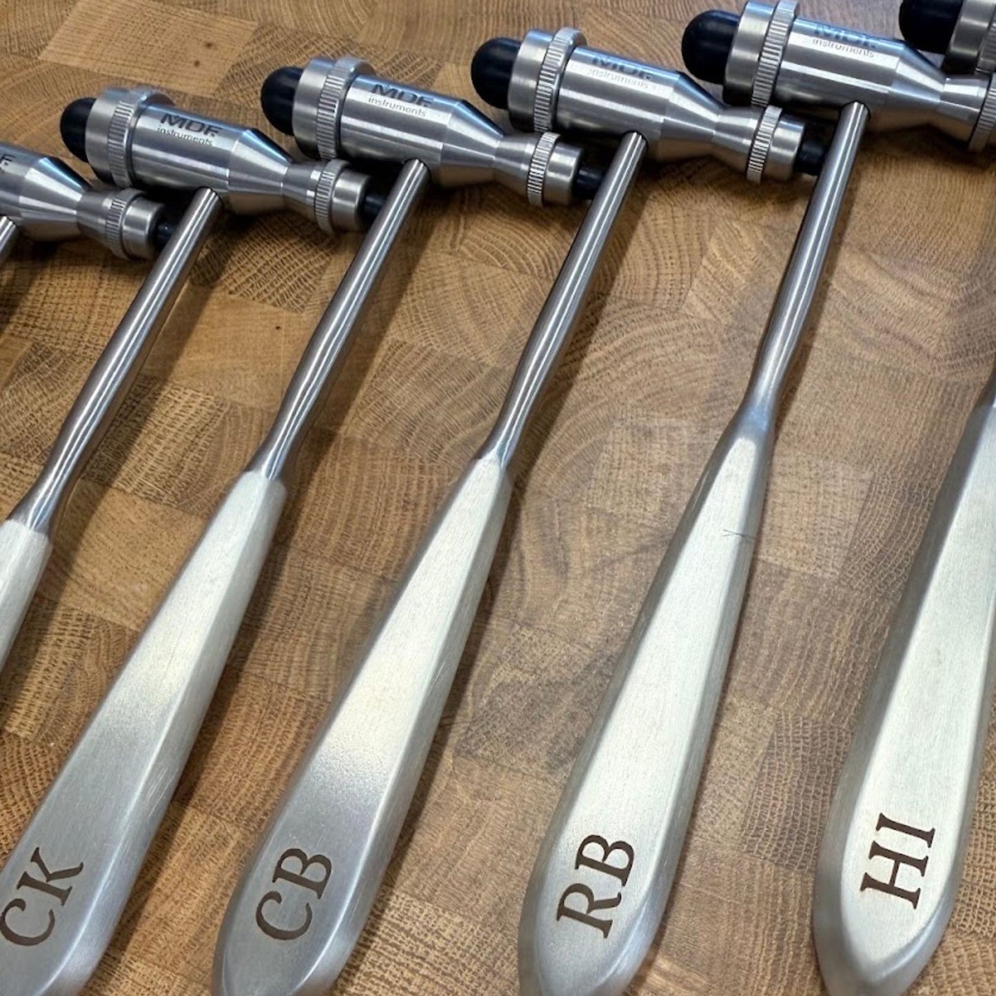 Four medical reflex hammers with engraved handles reading CK, CB, RB, and HI, arranged on a wooden surface.