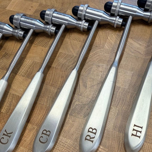 Four medical reflex hammers with engraved handles reading CK, CB, RB, and HI, arranged on a wooden surface.