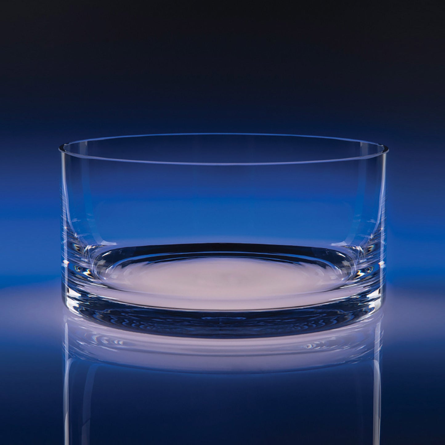 A clear glass bowl with a thick, sturdy base, shown on a reflective dark blue surface against a dark blue gradient background.
