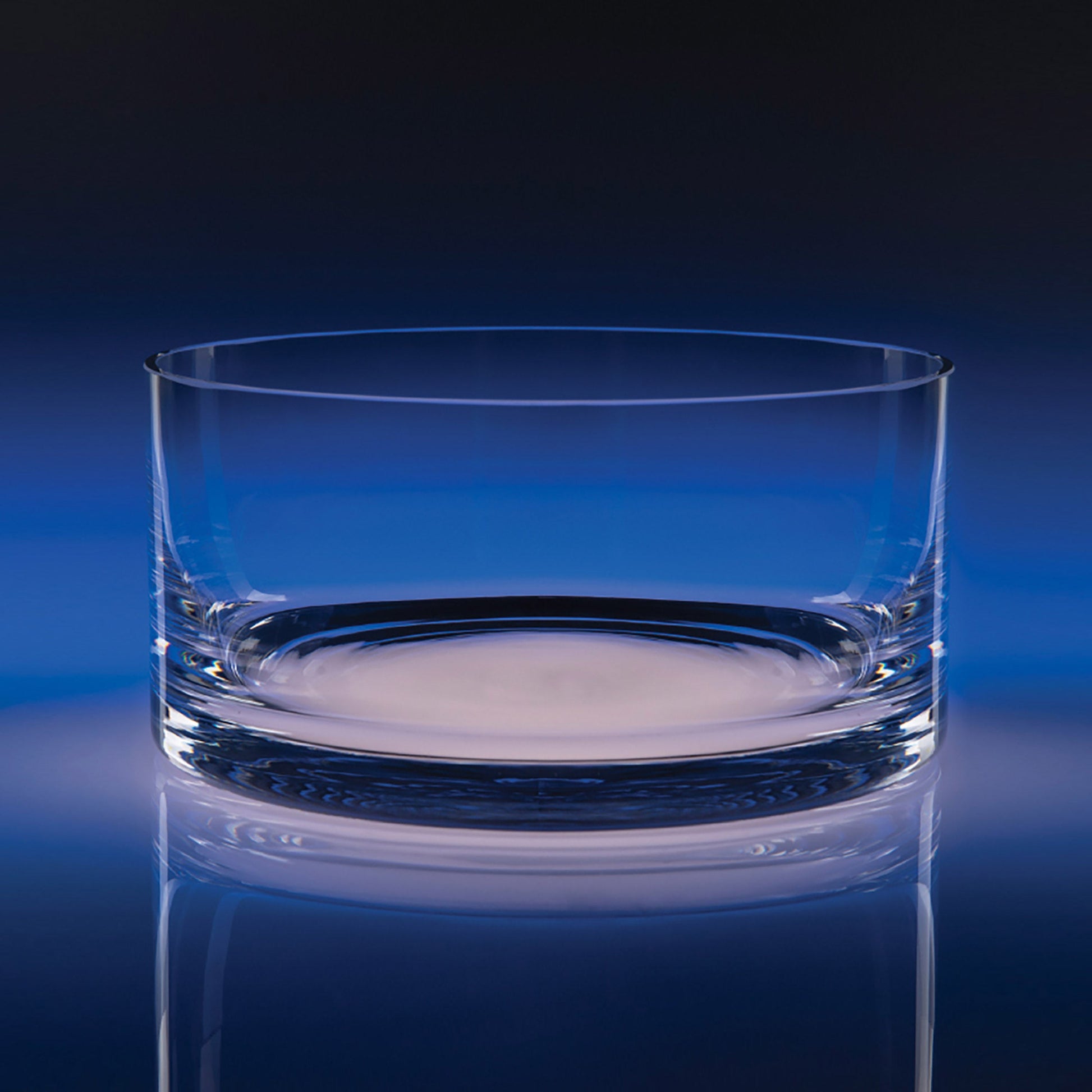 A clear glass bowl with a thick, sturdy base, shown on a reflective dark blue surface against a dark blue gradient background.