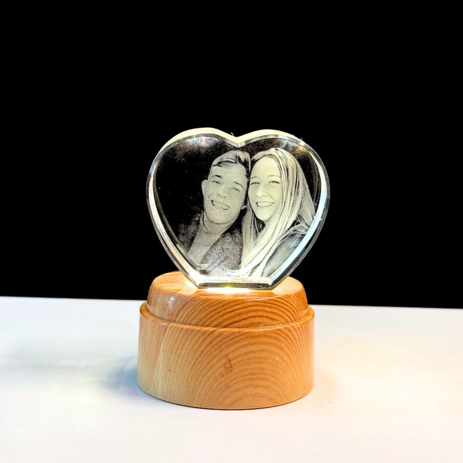 A heart-shaped clear glass keepsake photo plaque with an engraved black-and-white portrait of a couple, mounted on a round light wood base against a black background.