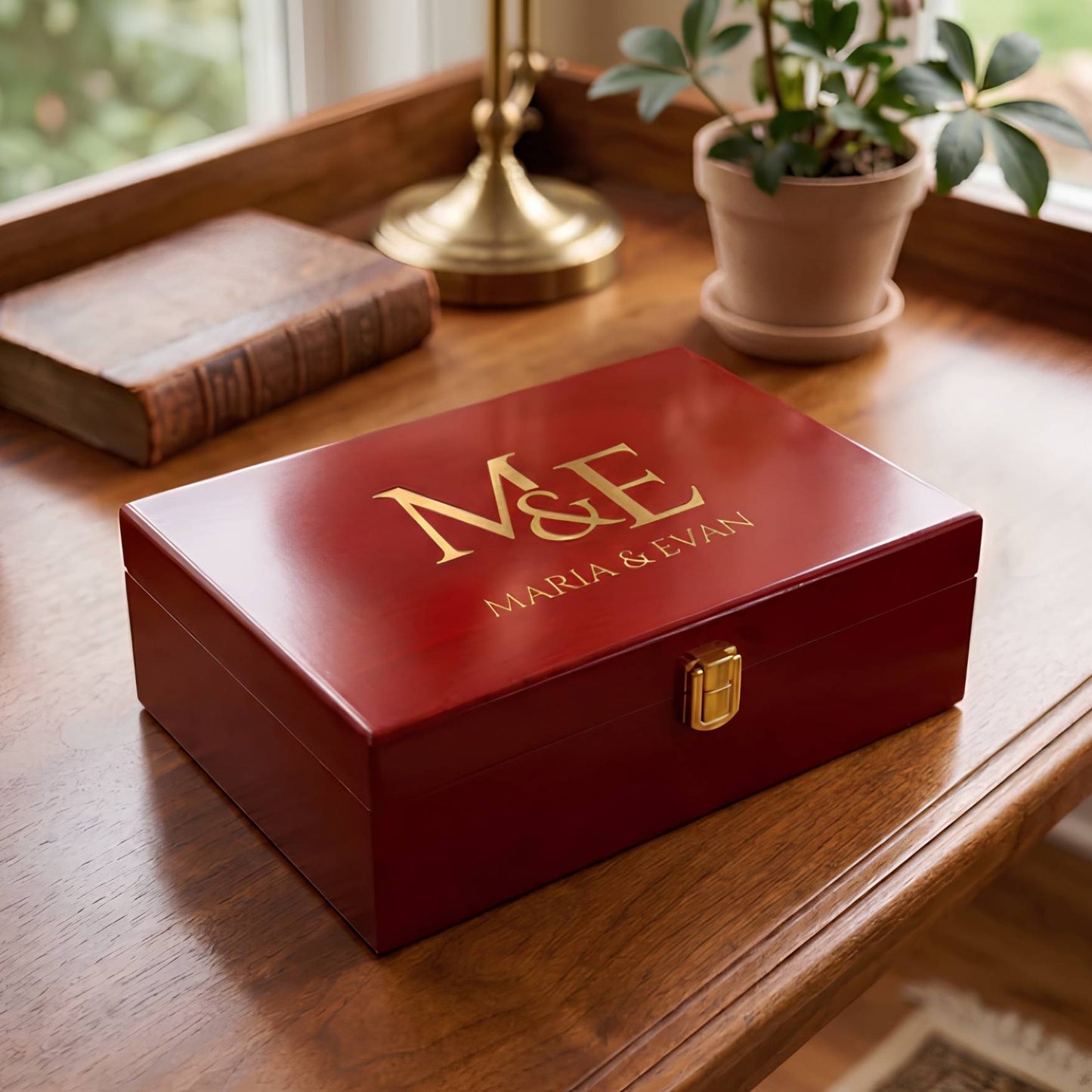 A personalized red wooden keepsake box with gold lettering reading 'M&E MARIA & EVAN' on the lid, placed on a wooden table next to a book and a potted plant.