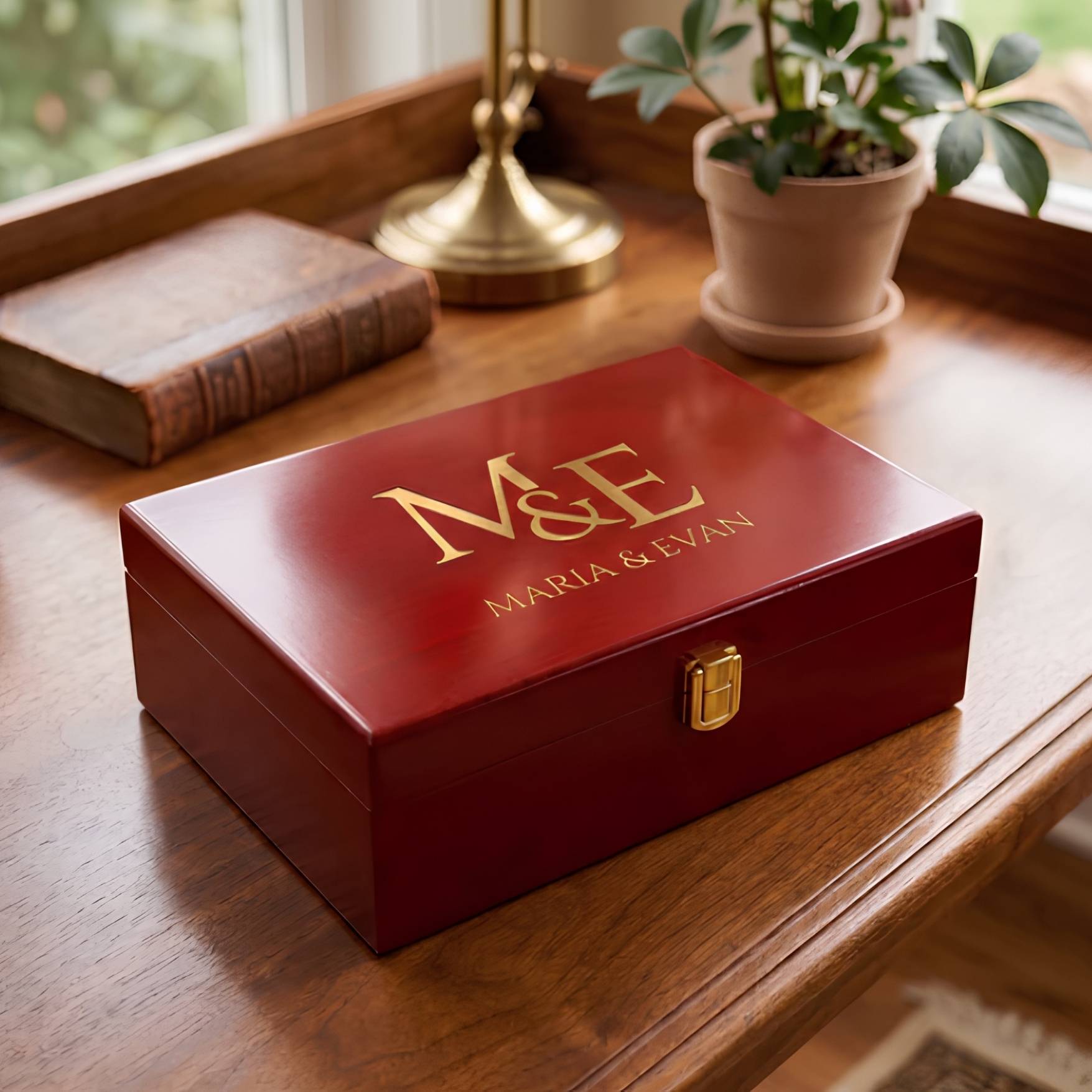 A personalized red wooden keepsake box with gold lettering reading 'M&E MARIA & EVAN' on the lid, placed on a wooden table next to a book and a potted plant.