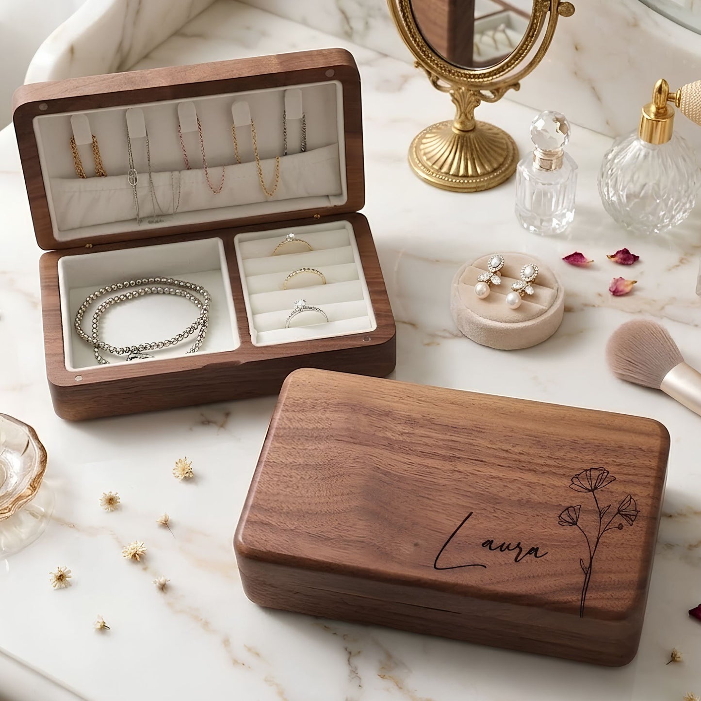 A personalized wooden jewelry box set, featuring an open box with velvet lining holding necklaces and rings, and its closed lid engraved with the name 'Laura' and a floral design, styled on a marble surface with cosmetics and gold accessories.
